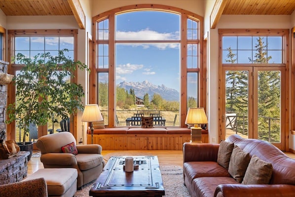 A cozy living room with a large window offering a mountain view, wooden furniture, and a fireplace. - Boundary JH - Three Peaks Cabin (Jackson)