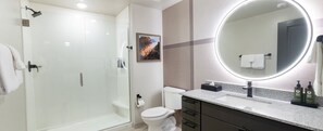 Ein modernes Badezimmer mit einer gläsernen Duschkabine, einer Toilette, einem Waschbecken mit Granitplatte und einem großen Spiegel mit runder Beleuchtung.
