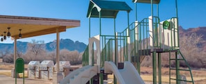 Ein Spielplatz mit Klettergerüsten und Rutschen, Grillmöglichkeiten und einem überdachten Picknickbereich mit Blick auf die Berge.