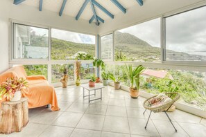 Ein geräumiger Balkon mit großen Fenstern, einem Korbstuhl, einem kleinen Tisch und verschiedenen topfgebundenen Pflanzen.