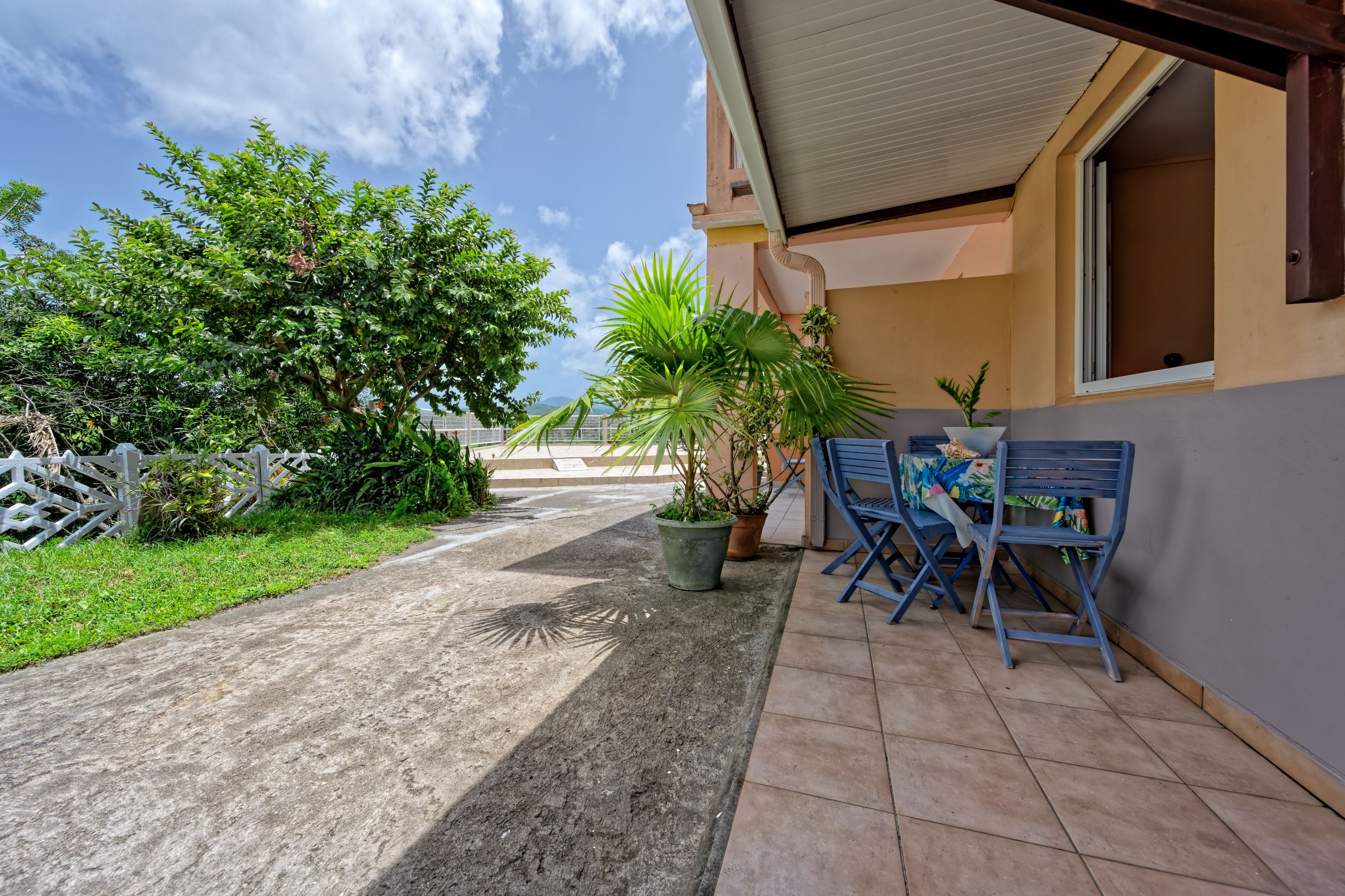 A paved pathway leading to a building with a covered outdoor seating area, potted plants, and a white fence.