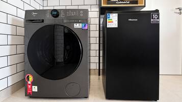Máquina de lavar e secar compacta com visor digital, frigorífico compacto e prateleira de madeira com máquina de café.