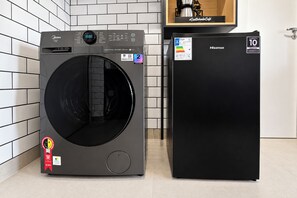 A compact washer and dryer unit with a digital display, a compact refrigerator, and a wooden shelf with a coffee maker. - Stay Home Itaim - Piscina - Academia (São Paulo)