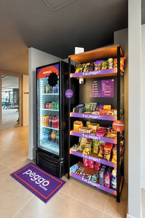 A vending machine stocked with beverages and snacks, with a purple mat on the floor and a sign indicating payment options. - Stay Home Itaim - Piscina - Academia (São Paulo)