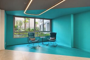 A room with teal walls, two chairs, a small table, and a large window with a view of greenery and buildings. - Stay Home Itaim - Piscina - Academia (São Paulo)