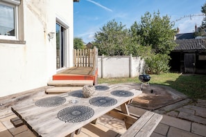A wooden table with black circular coasters, a barbecue grill, and a wooden bench with steps leading to a door. - L'Endurance - Proche 24h du Mans (Le Mans)