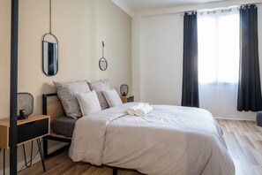 A neatly made bed with a white duvet and pillows, a bedside table with a lamp, and a large window with curtains. - Le Relais - Proche 24h du Mans - Bus (Le Mans)