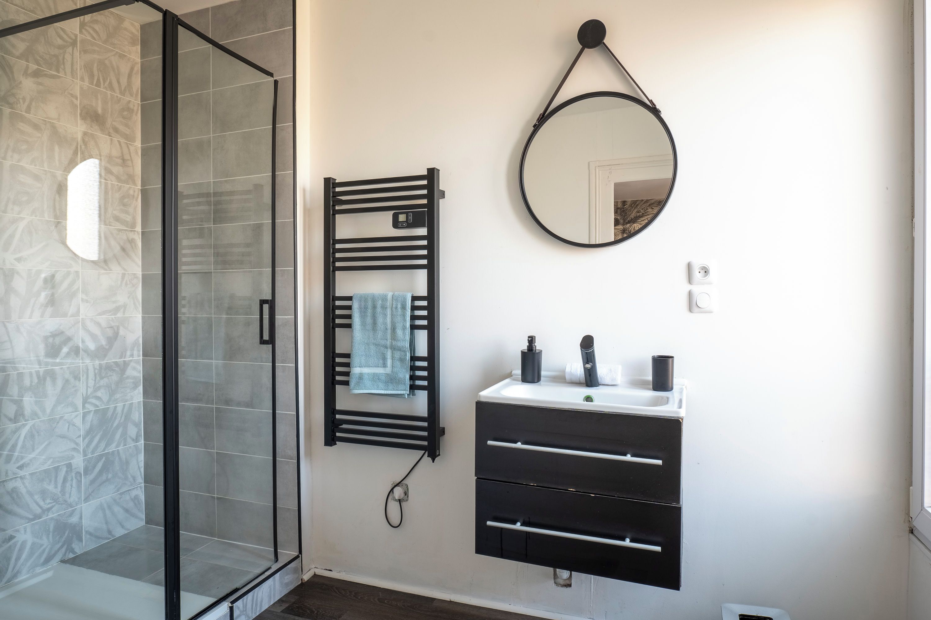 A bathroom with a glass shower enclosure, a towel rack with a towel, a mirror, and a sink with a faucet and soap dispenser.