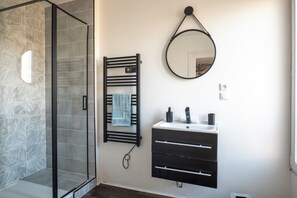 Ein Badezimmer mit einer Duschkabine aus Glas, einem Handtuchhalter mit Handtuch, einem Spiegel und einem Waschbecken mit Wasserhahn und Seifenspender.