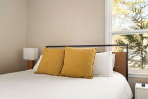 A bed with white bedding and two mustard-colored pillows, a bedside table with a lamp, and a window with a view of trees. - 13 Guests & Fire Pit, Double Living, 2026 Refresh (Raleigh)