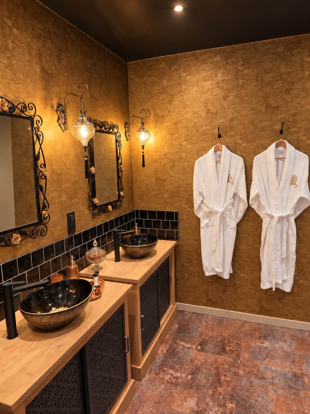 A bathroom with a wooden vanity, black and gold tiles, a large mirror, wall-mounted lights, and two white robes hanging on hooks.