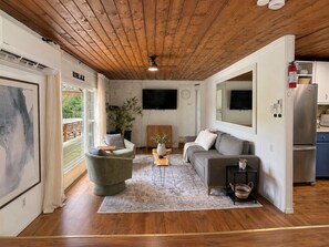 A modern living room with a wooden ceiling, a grey sofa, a green armchair, a coffee table, and a large window. - Cozy Lakefront Cottage with Fireplace | ATV & Snowmobile Getaway near Bancroft | Sleeps 6 (Faraday)
