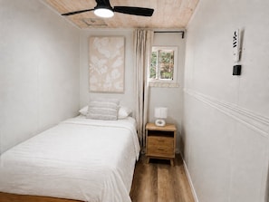 Une chambre à coucher avec un lit, un ventilateur de plafond, une fenêtre avec des rideaux, une table de chevet avec une lampe et un interrupteur fixé au mur.