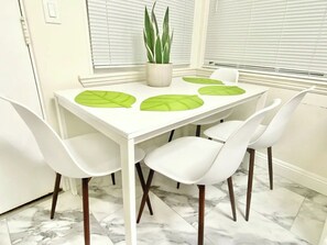 Taula de dinar amb quatre cadires blanques, una planta en test i estores verdes.