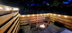 Ein Außenbereich mit Holzbänken und -stühlen, einer Feuerstelle und Lichterketten.