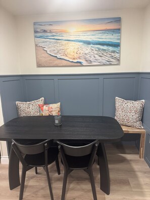 A dining area with a dark wooden table, two black chairs, a bench with patterned cushions, and a wall-mounted painting of a beach scene. - Surfcity Getaway 5min walk to beach and Main St (Huntington Beach)