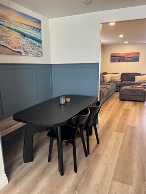 A dining area with a dark wooden table and chairs, a painting of a sunset on the wall, and a view into a living room with a couch and a painting of a pier. - Surfcity Getaway 5min walk to beach and Main St (Huntington Beach)