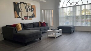 A modern living room with a sectional sofa, a coffee table, and a large window with blinds. - Family-Friendly 3BR/2BA Condo Minutes from Disney with Pool & Gym (Kissimmee)