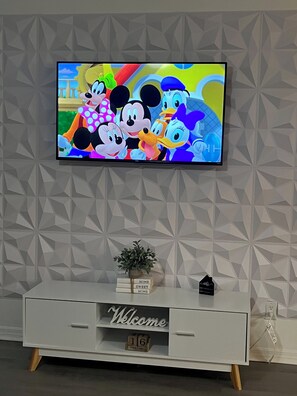 A television mounted on a wall with a geometric pattern, a white console table with a welcome sign, and a small plant. - Family-Friendly 3BR/2BA Condo Minutes from Disney with Pool & Gym (Kissimmee)