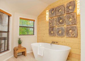A bathroom with a freestanding bathtub, wooden wall decor, a window with blinds, and a small wooden stool with a plant. - Still Waters Resort 1BR Condo – Pools, Lake & Marina (Branson)