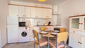 Een kleine keuken met een wasmachine, magnetron en een eethoek met stoelen en een tafel.