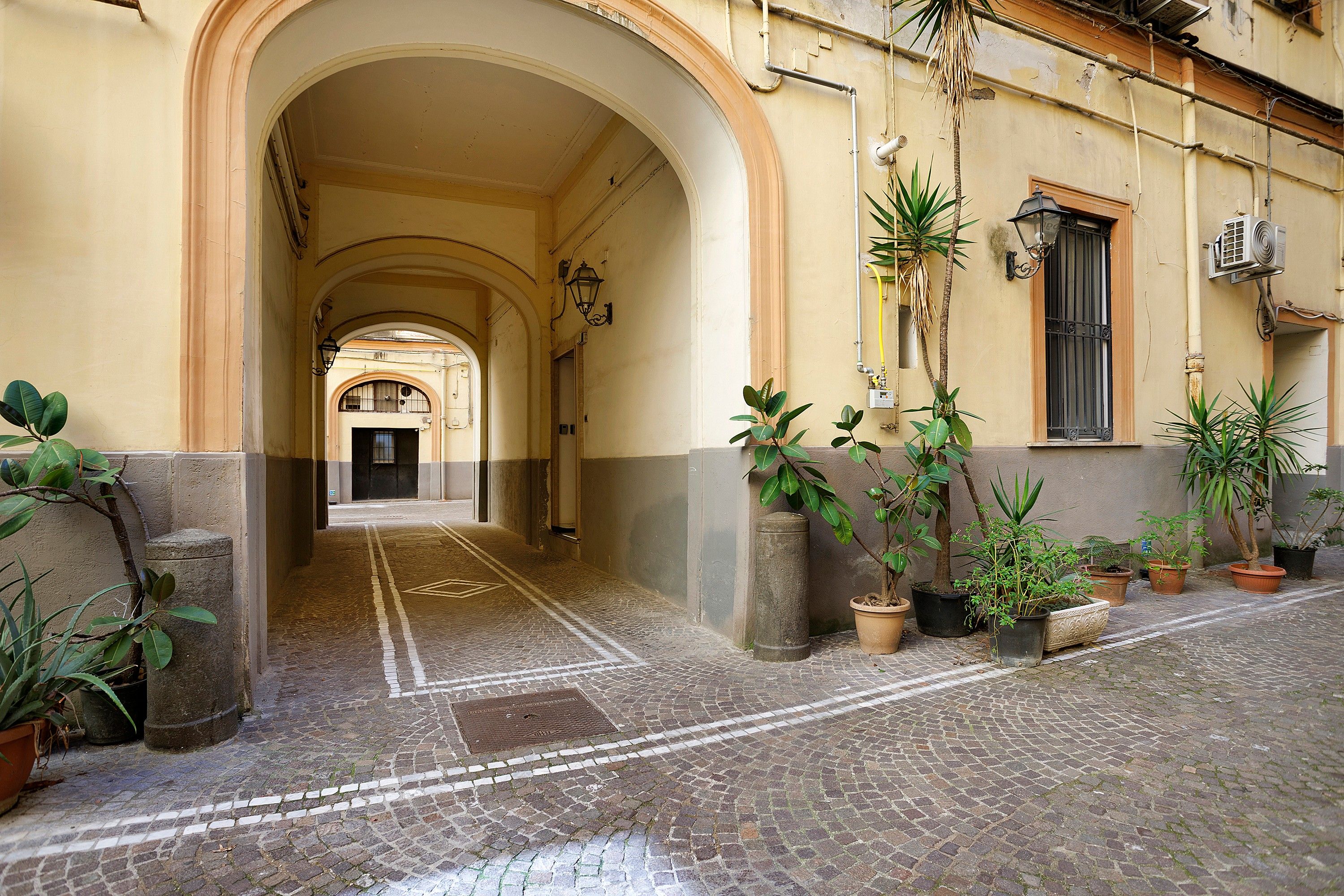 A cobblestone street with an arched entrance leading to a building with multiple windows and a door. There are potted plants along the side of the street.