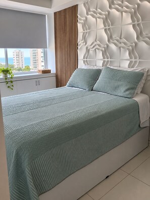 A modern bedroom with a large bed, a window with a view of buildings, and a decorative wall panel. - Apartment, Ocean View (Cartagena de Indias)