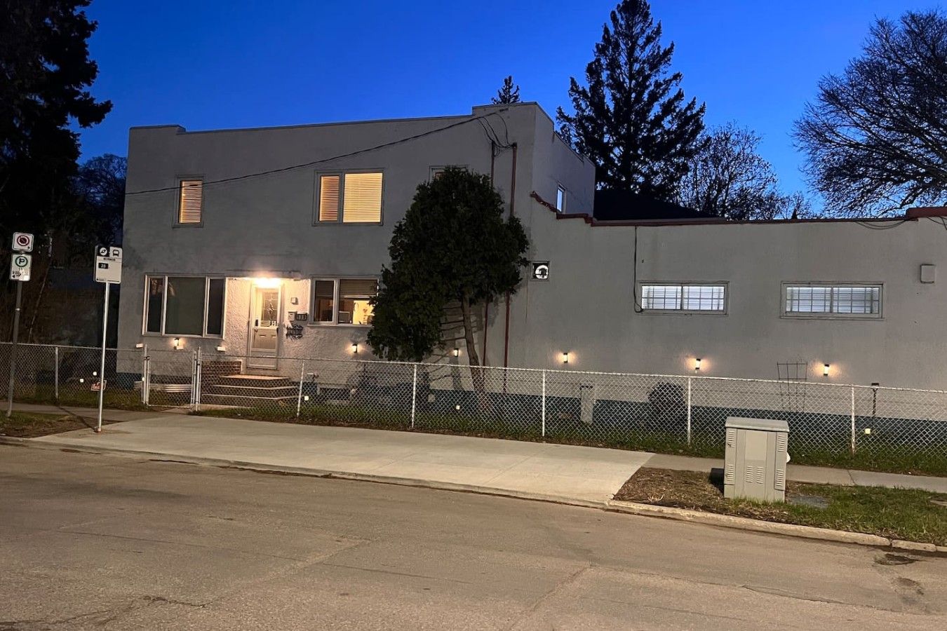 A two-story building with a fenced front yard, a visible entrance, and a street sign indicating parking restrictions.