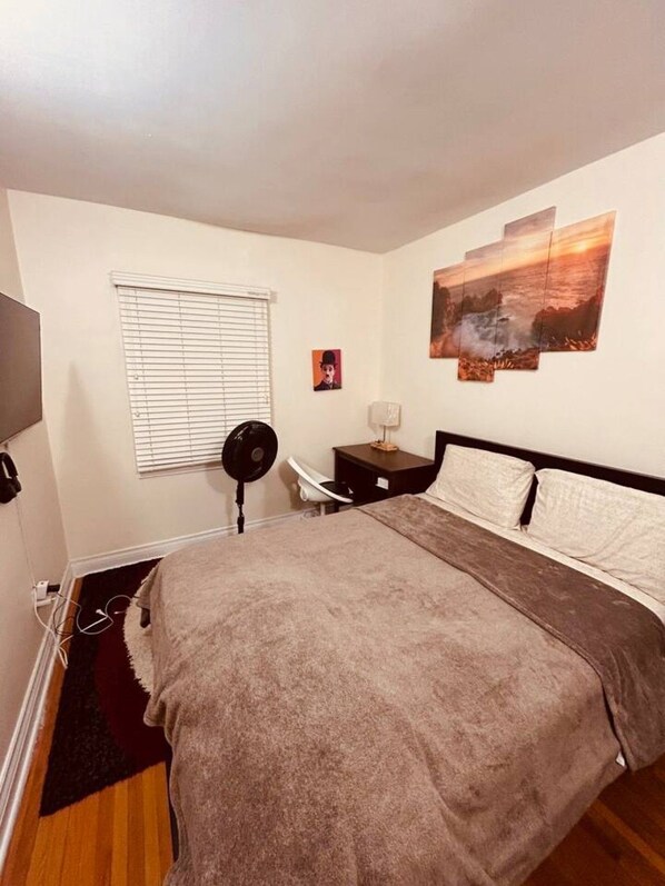 Un dormitorio con cama, mesita de noche, lámpara, ventana con persianas y un cuadro colgado en la pared.