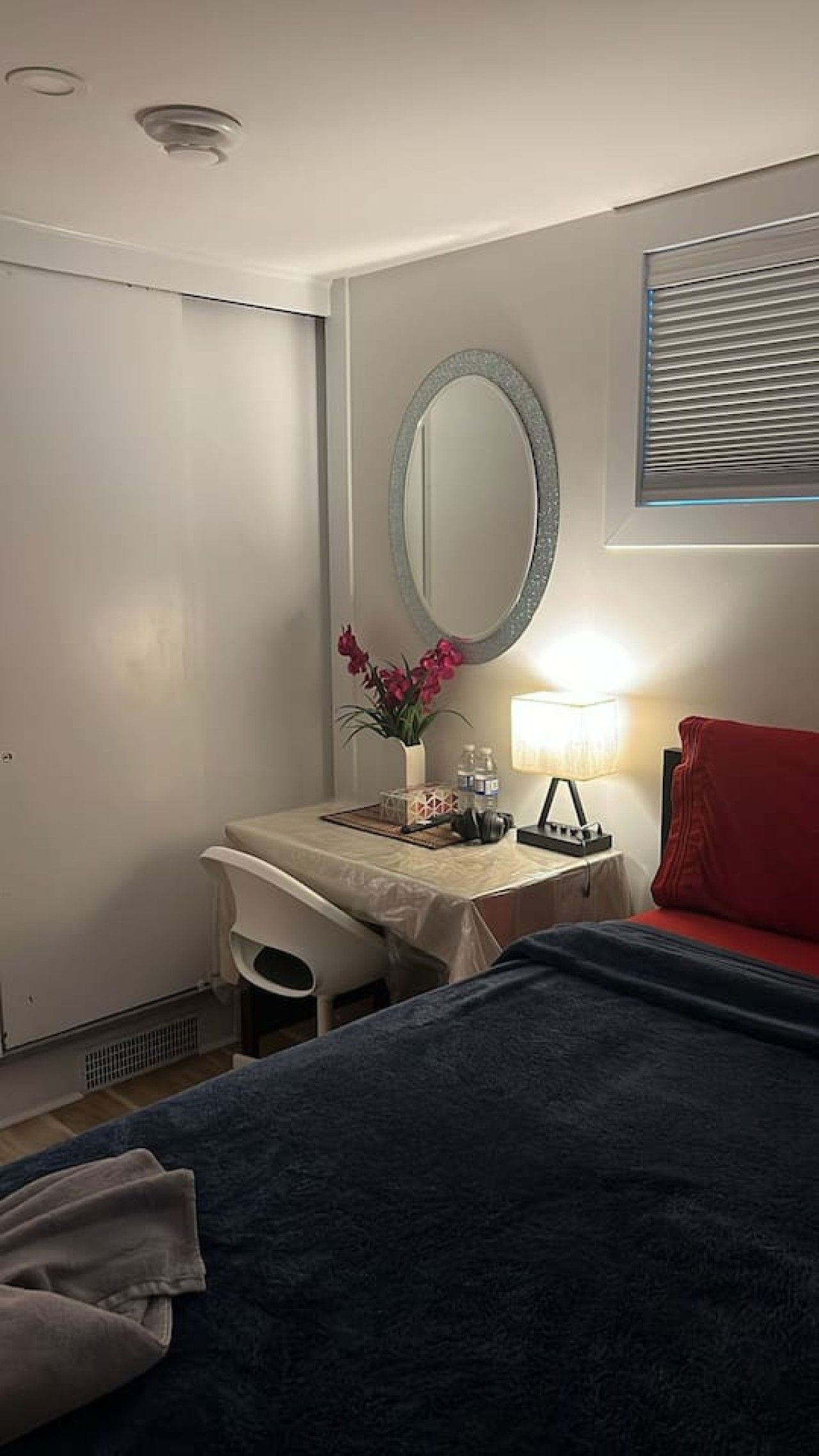 A hotel room with a bed, a desk with a chair, a mirror, a lamp, and a window with blinds.