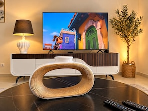 A modern living room with a flat-screen TV, a decorative woven stool, a potted plant, and a remote control on a black table. - Sea View Apartment w/ Pool & Parking in Canico (Caniço)