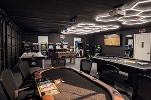 Una mesa de billar con taco, sillas y una mesa de futbolín en una sala con juegos de arcade y una lámpara de techo neón.