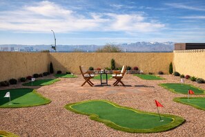 Un campo de mini golf con una mesa y sillas en el centro, rodeado por un jardín ajardinado y vistas a montañas al fondo.
