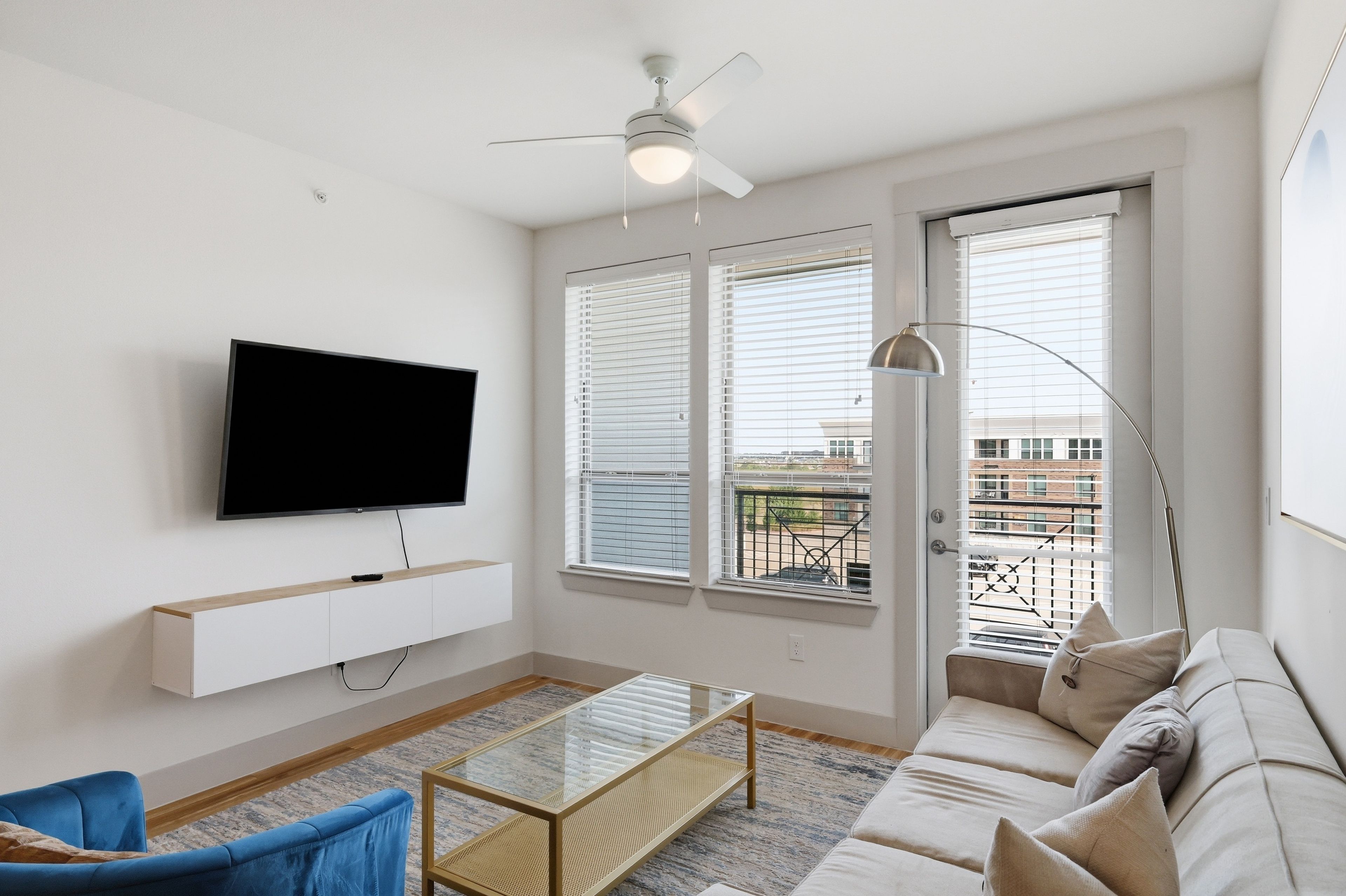 Una sala de estar moderna con un televisor de pantalla plana montado en una pared blanca, un ventilador de techo y un gran ventanal con persianas.