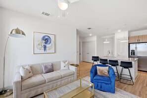 Ein modernes Wohnzimmer mit einer Couch, einem blauen Sessel, einem Couchtisch und einer Stehlampe. Es gibt eine Küchenzeile mit einem Kühlschrank, einer Spüle und Barhocker.
