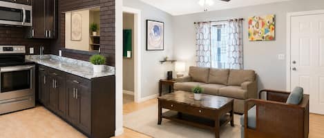 A modern kitchen with dark cabinetry and a stainless steel microwave, a living area with a sofa and coffee table, and a ceiling fan.
