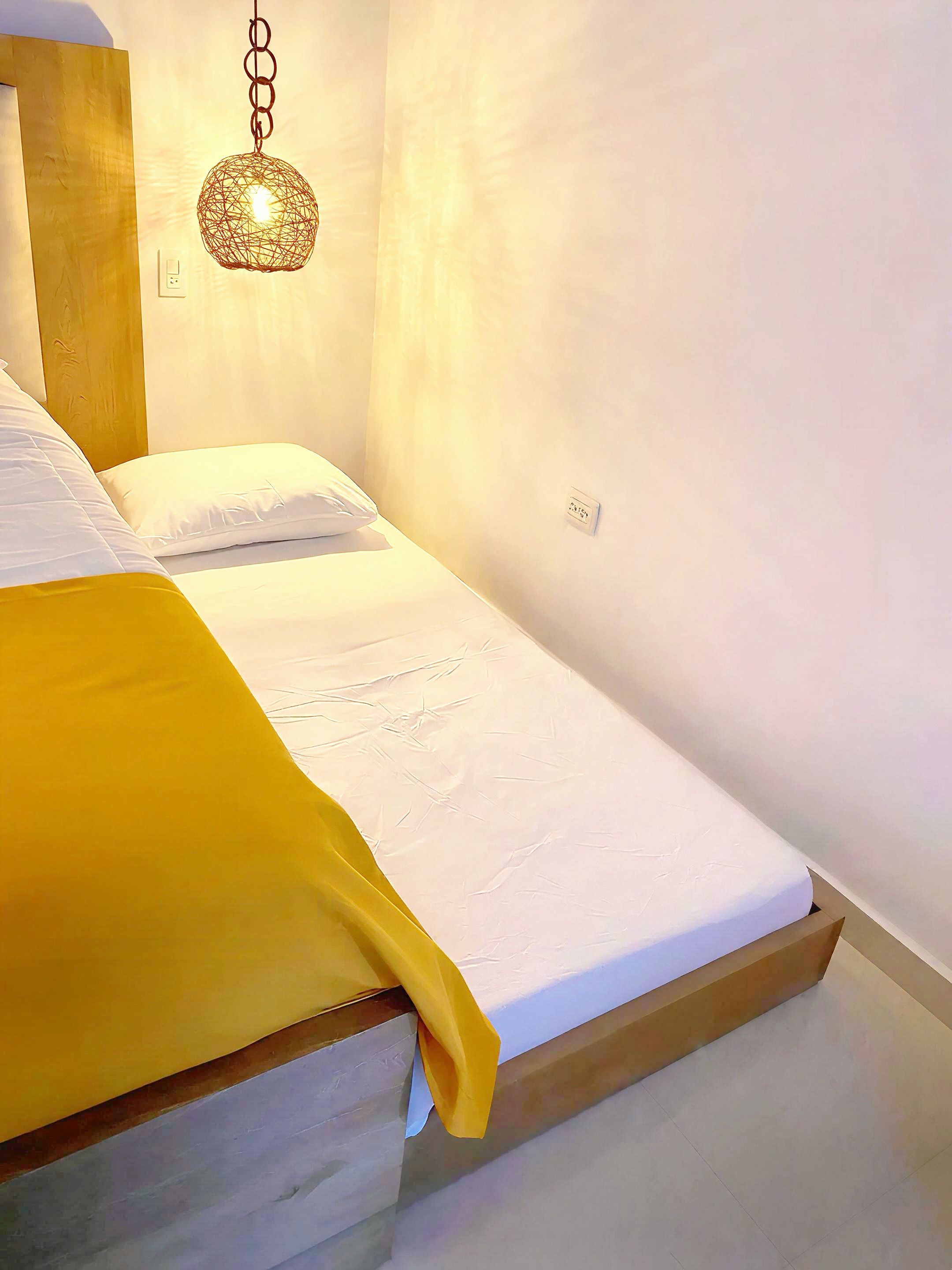 A single bed with a white mattress and a yellow blanket, a wooden headboard, and a hanging light fixture.