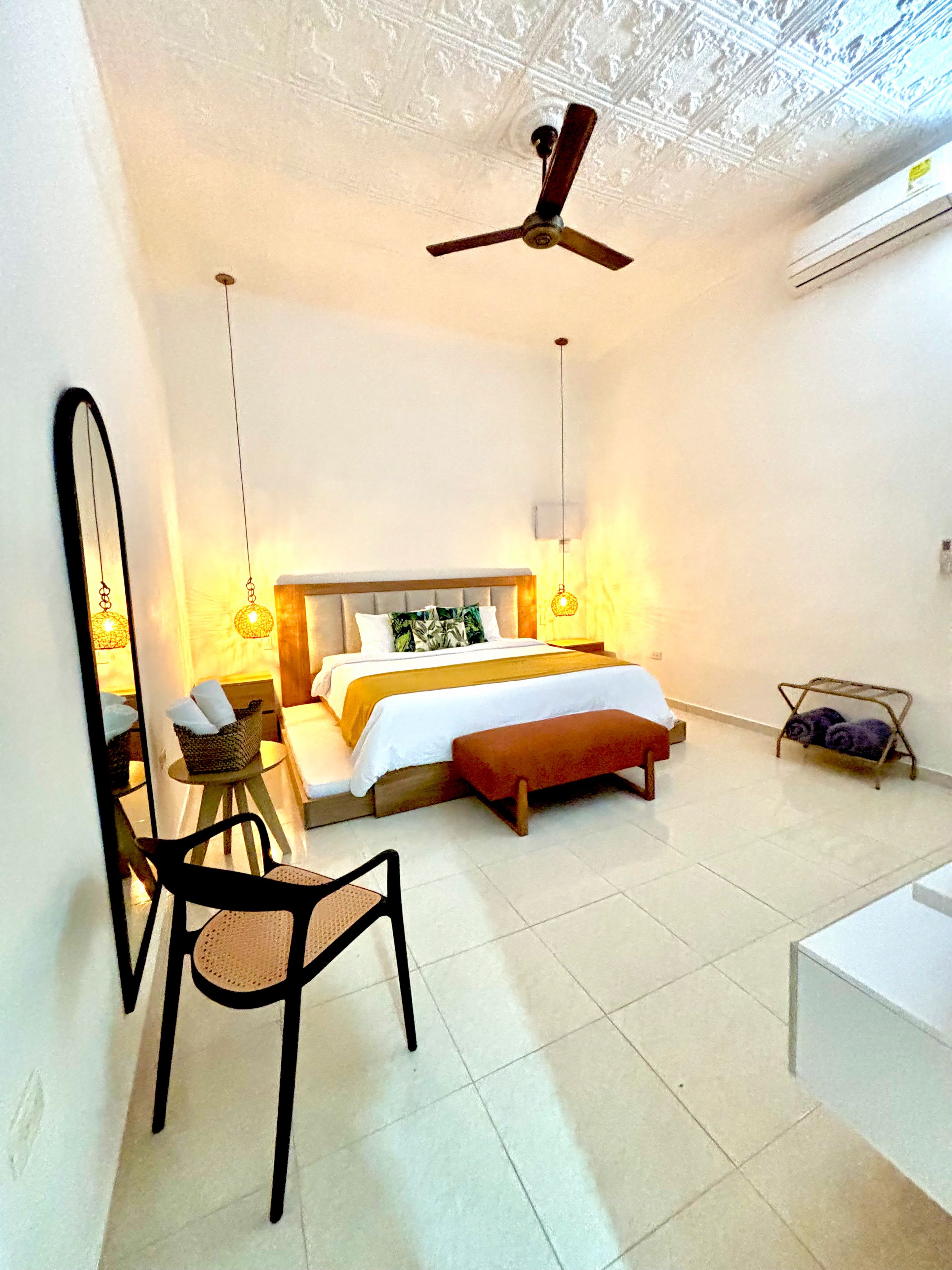 Quarto com cama, cadeira, mesa de cabeceira, luminária pendente e ventilador de teto.