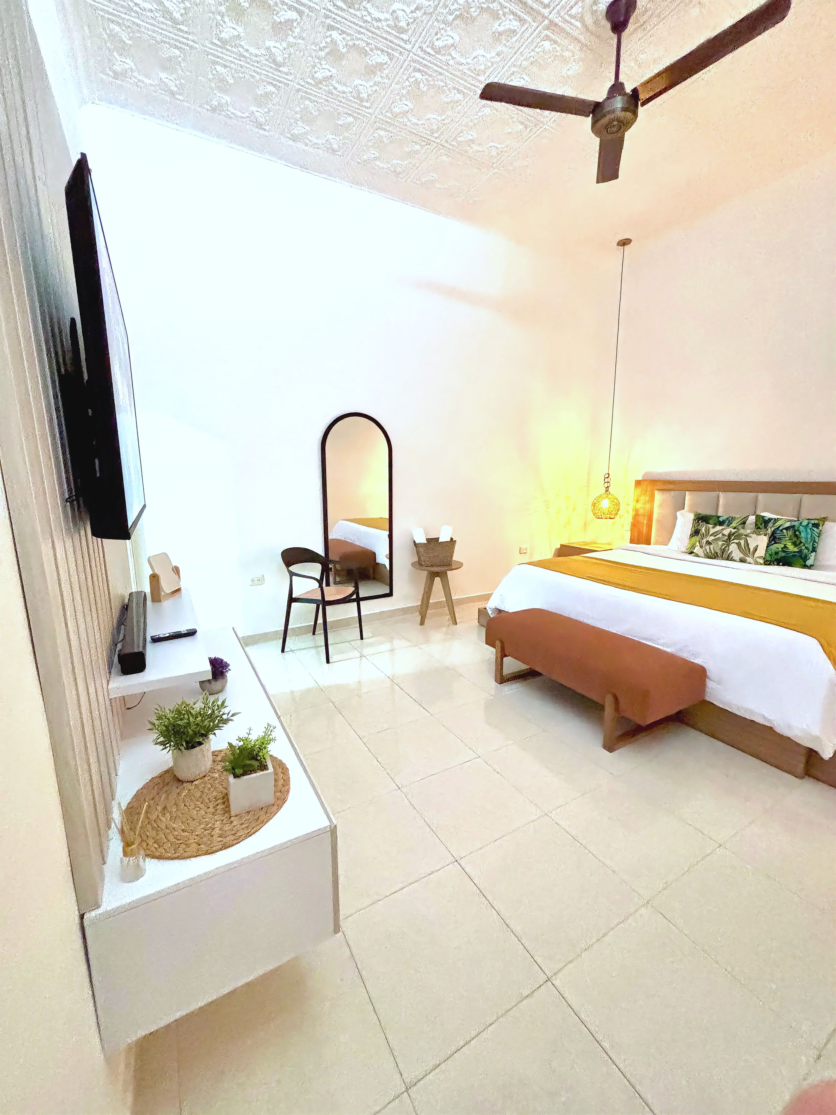 Quarto de hotel com cama, escrivaninha, cadeira, televisão e ventoinha no teto.