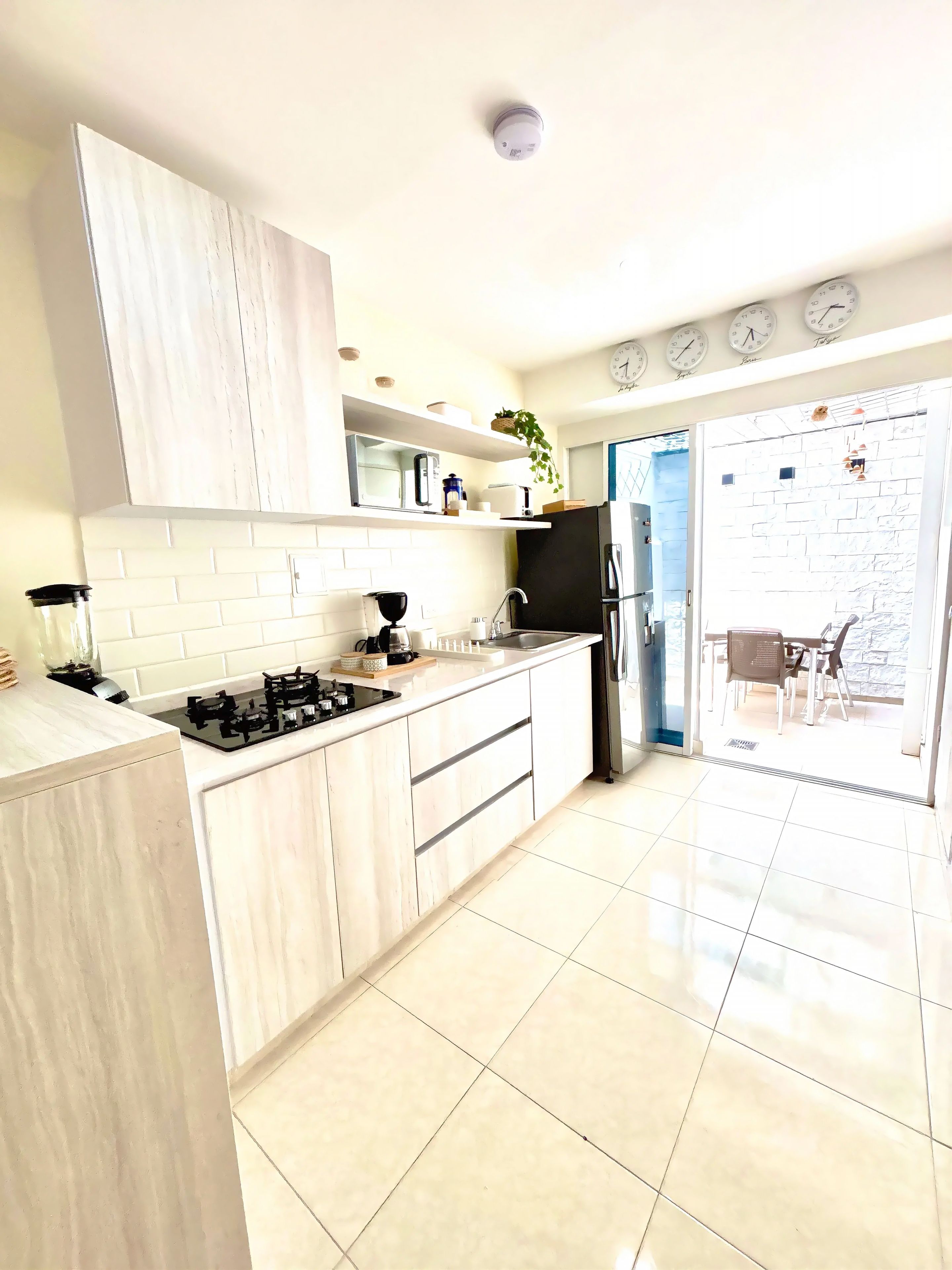 A modern kitchen with white cabinetry, a black stove, and a refrigerator. There are clocks on the wall and a plant on the counter.