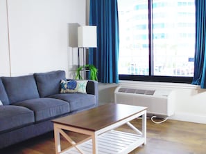 Ein Wohnzimmer mit einer Couch, einem Couchtisch, einer Stehlampe, einem Blumentopf und einem Fenster mit Vorhängen.