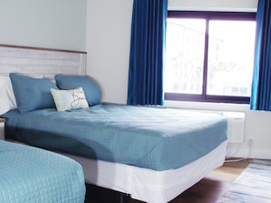 Ein Bett mit blauer Bettdecke und Kissen, ein Fenster mit blauen Vorhängen und ein Blick auf Gebäude im Freien.