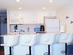 Una cocina moderna con armarios blancos, microondas, cafetera y refrigerador. Hay tres taburetes blancos con patas negras.
