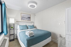 Ein ordentlich eingerichtetes Schlafzimmer mit Bett, einem Fenster mit Vorhängen, einem Heizkörper, einem Wandbild und einer Deckenleuchte.