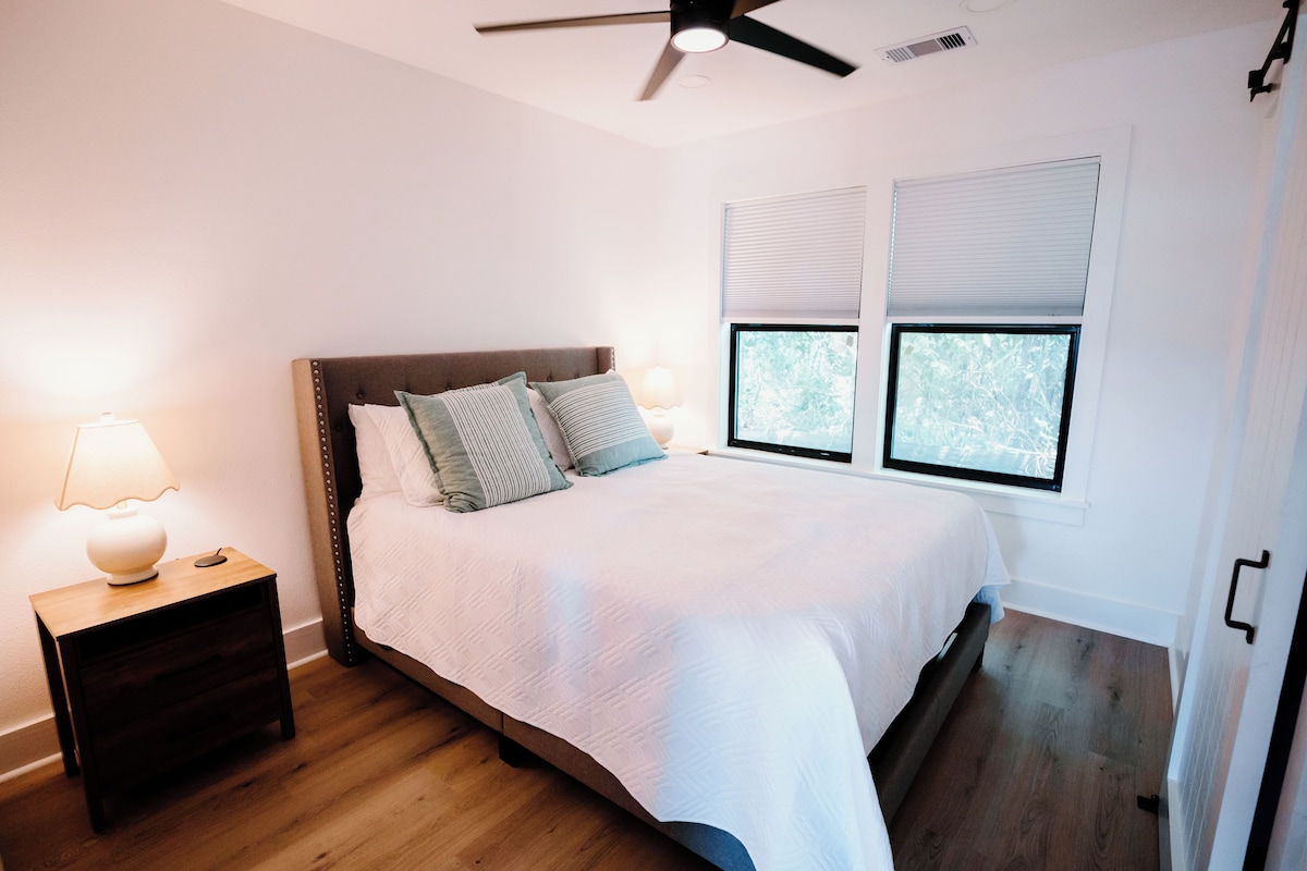 A bedroom with a bed, nightstand, lamp, ceiling fan, and windows.