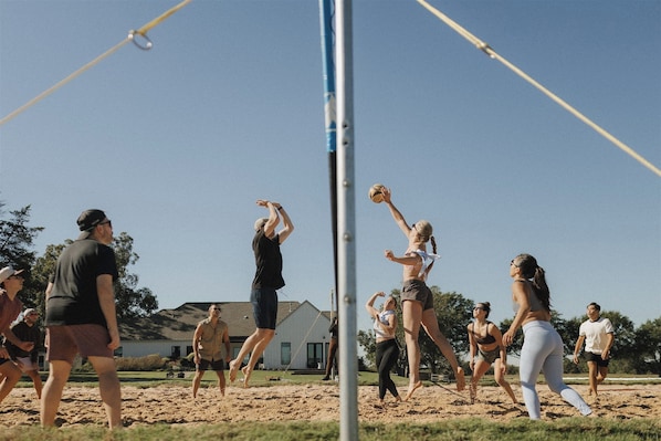 A group of people playing volleyball on a sandy court with a house in the background.