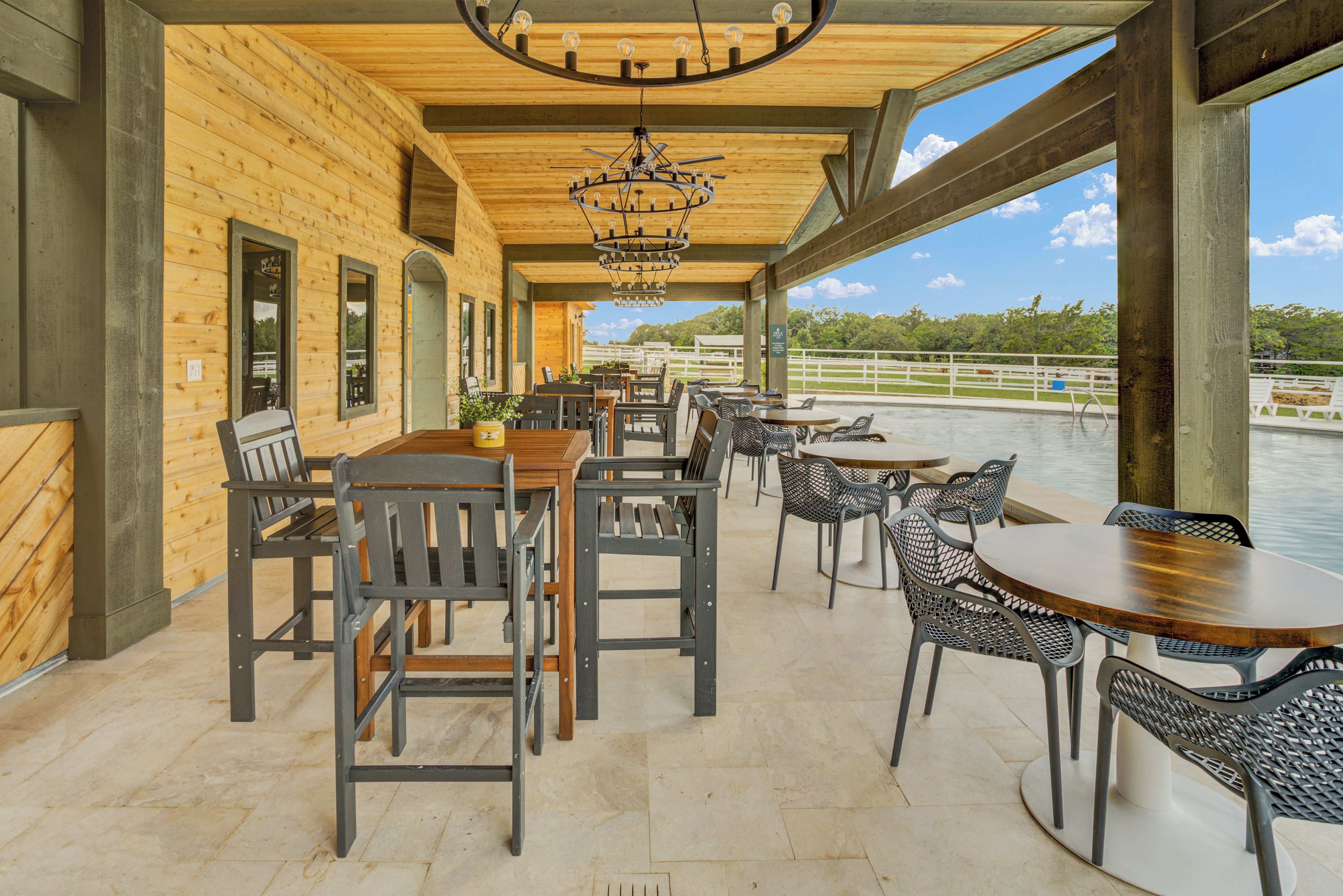 An outdoor dining area with wooden tables and chairs, a chandelier, and a view of a body of water.