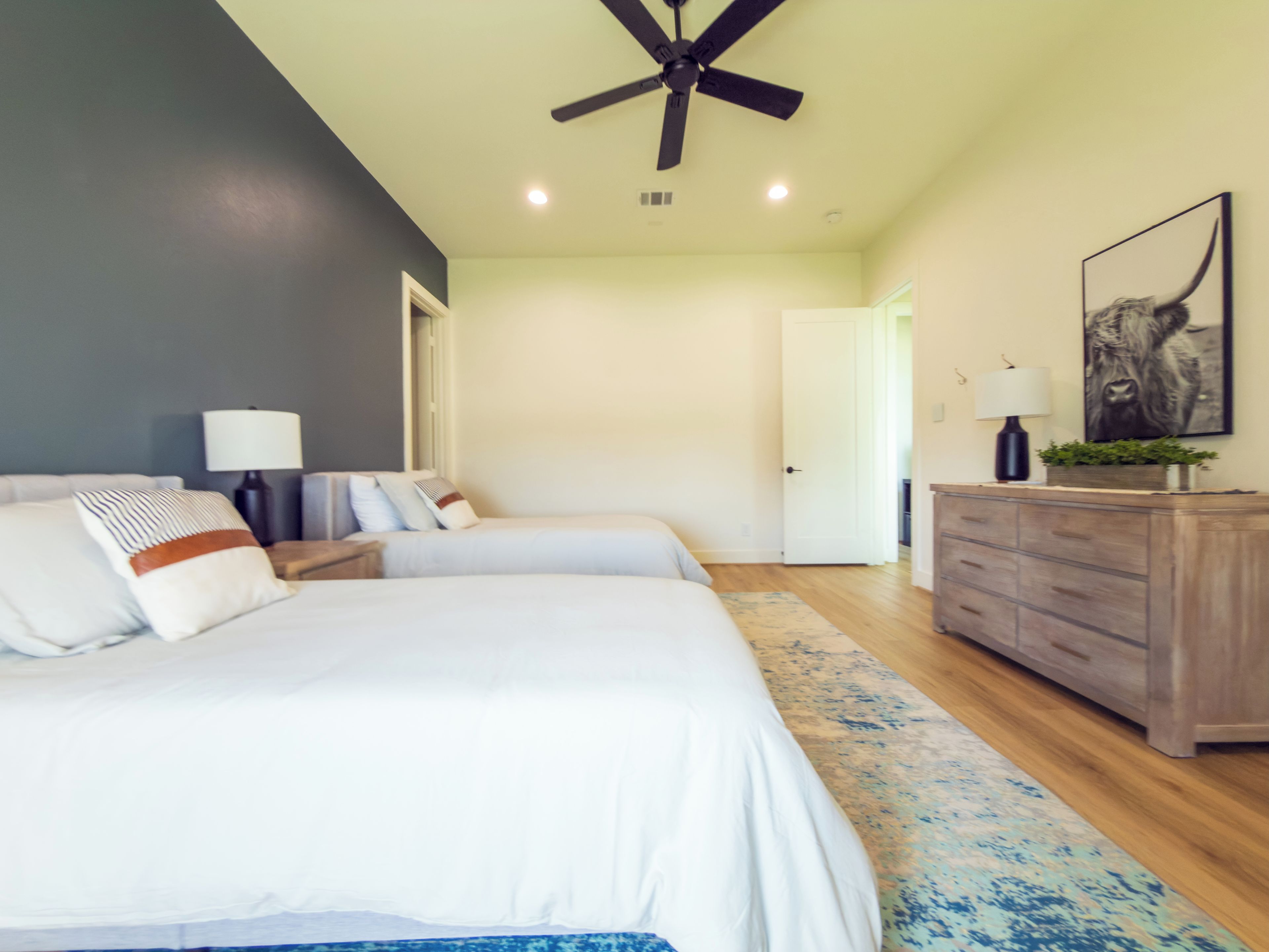 A bedroom with two beds, a ceiling fan, a nightstand with a lamp, a dresser, and a door leading to another room.