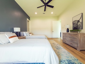 A bedroom with two beds, a ceiling fan, a nightstand with a lamp, a dresser, and a door leading to another room.