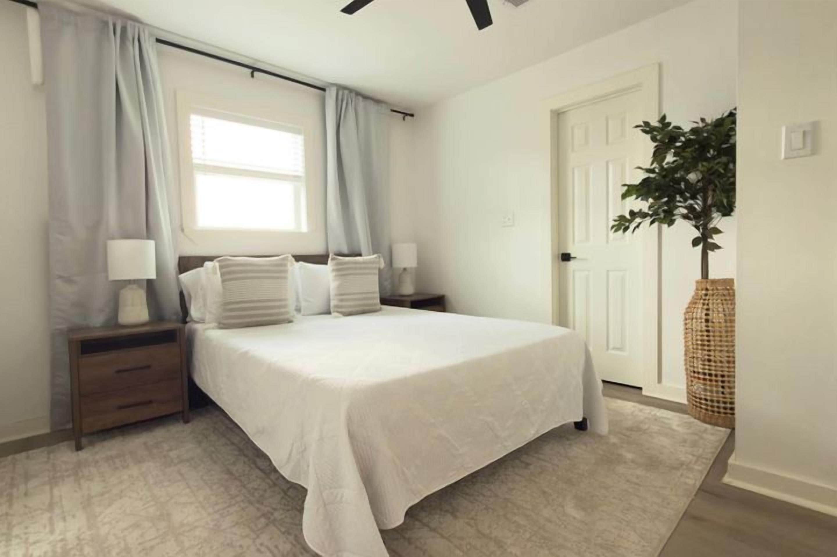 A bedroom with a bed, nightstands, a window with curtains, a door, and a plant.